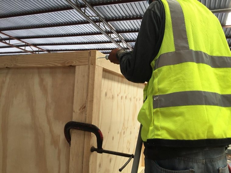 Trabajador con chaleco reflectante junto a una caja de madera en un área cubierta.