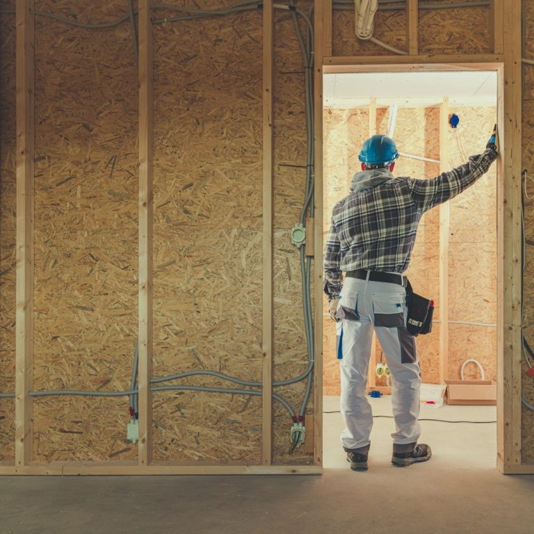 Hombre con casco y herramientas en una construcción, recargado sobre el marco de la puerta, observando hacia una habitación.