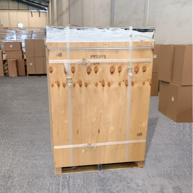 Caja de madera sin abrir, con parte de envolvente plástico y cinta de embalaje, en un almacén.