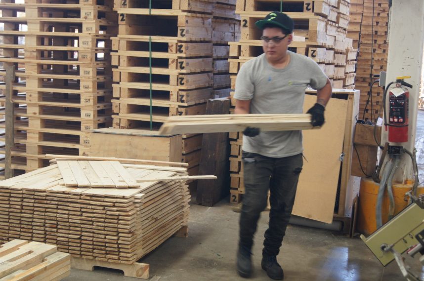 Operario de Prommont en proceso de ensamble de armazones Hombre joven en un taller sosteniendo varias tablas de madera durante proceso de ensamblado de armazones, rodeado tarimas de madera.